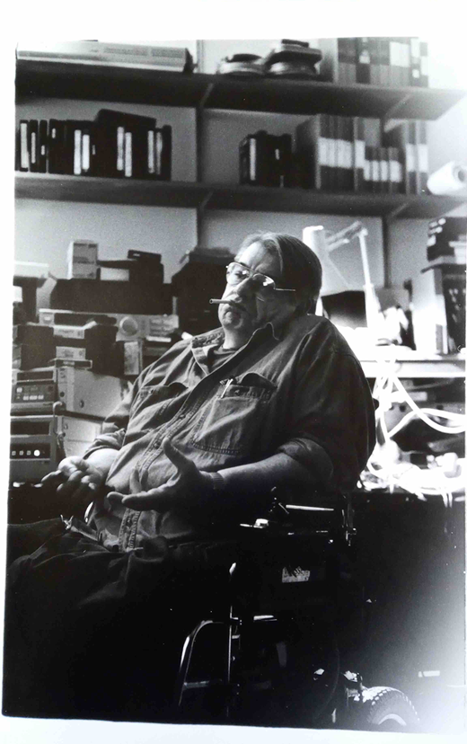 A light-skinned man in his fifties sits in his electric wheelchair in the foreground. Dressed in a casual shirt with rolled-up sleeves, he has an unlit cigarette in his mouth. The light reflected in his glasses partially obscures his eyes which look outward away from the camera. He looks contemplative and his hands are gesturing as if in conversation. Behind him in his studio is an assortment of audio-visual equipment, cables, computers and an angled lamp on a series of tables. On the shelves behind the studio tables there are VHS tapes, folders, and film canisters lined up in an orderly fashion.