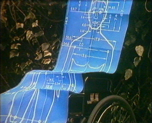 A grainy VHS image shows a manual wheelchair with large wheels and hand rims, mostly obscured by a large full-sized blue roll of paper which depicts a series of measurements across an image, printed in white, of a two -dimensional male figure. The folded and unfolded paper is draped over the wheelchair in a way that demonstrates how poorly the wheelchair can accommodate the figure of a man. In the background behind the chair and the paper image of the man are pale ivy leaves and other plants.