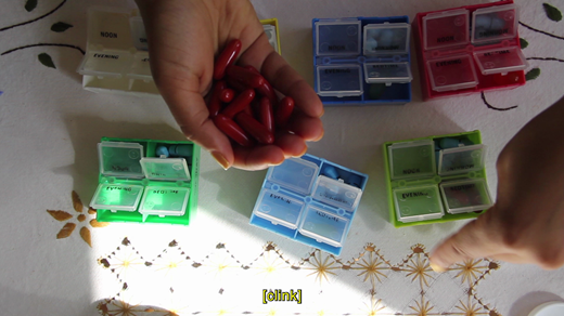 A still image from Lazard’s Crip Time depicting a brown human hand with yellow nail polish holding red pills above seven pill organizers in different colors. The open pill bottles are placed on an embroidered tablecloth at the bottom of the frame, while a second hand is approaching one of them. The word “clink” in parentheses appears as a yellow caption at the bottom of the screen.
