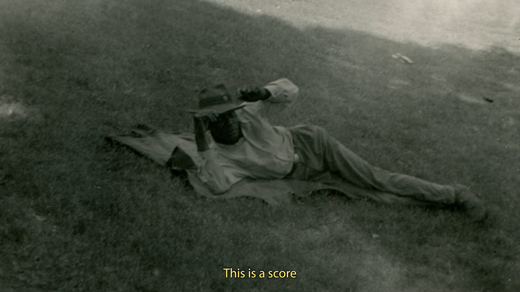 Historical footage of a Black man wearing a brimmed hat and reclining on a blanket in the grass is captioned in yellow, “This is a score.”