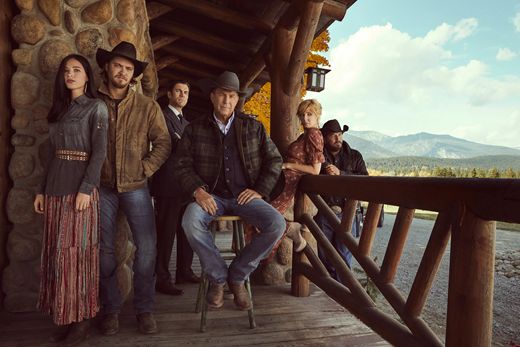 A large family portrait style photo of six people who all appear to be white. The man in the center, who appears older than everyone else, is seated on a stool and wearing a cowboy hat. Standing slightly behind him is a man wearing a suit. He is the only man in the photo not wearing a cowboy hat. On each side of the seated man there is a couple standing (a man and a woman). They are posed on the wooden porch of a stone house, overlooking a mountain landscape.