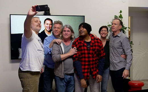 A large photograph of a group of smiling middle-aged people standing together taking a selfie. The group is comprised of seven men and one woman; one of the men appears virtually on a large screen behind the group.