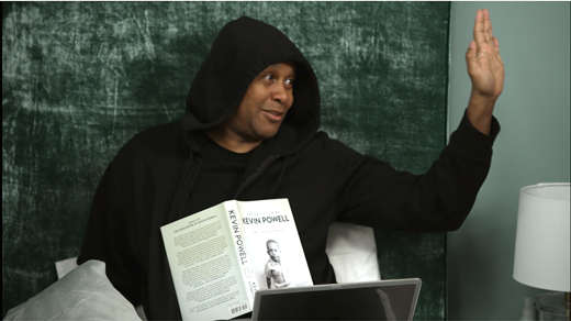 A large image shows a Black man in a black hooded sweatshirt. He is seated on a bed with his back pushed up against the headrest. He is smiling and looking off to one side, with an open book on his chest. The title of the book is Kevin Powell.