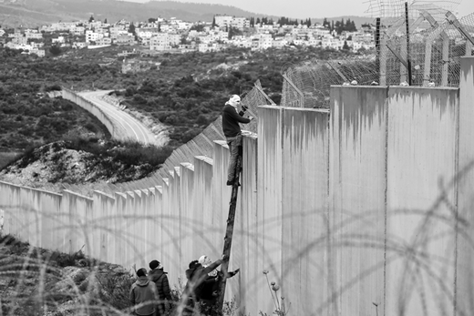 A masked Palestinian in Bil‘in, a village near Ramallah, cuts through the barbed wire lining the separation wall with Israel. In the background is Mod'in Ilit, one of the largest Israeli settlements in the West Bank. (7 February, Abbas Momani/AFP via Getty Images)